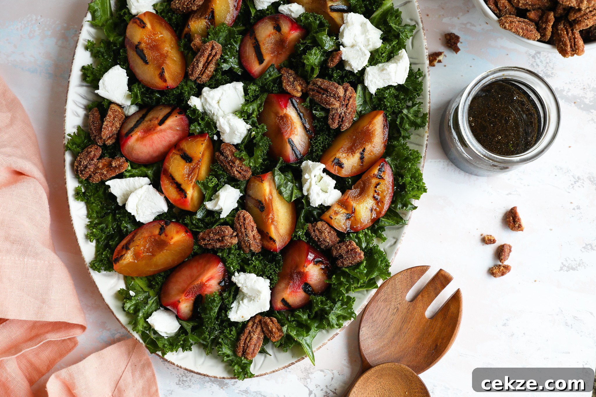 A grilled stone fruit kale salad surrounded by a jar of maple balsamic dressing, wooden salad tongs and a pink linen napkin