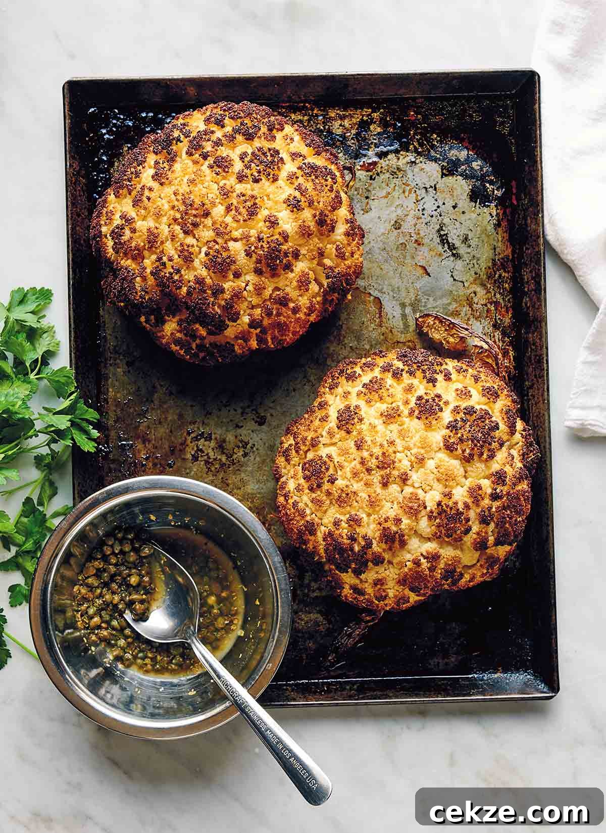 Crispy Spicy Roasted Cauliflower with Chili and Garlic 9 Two whole roasted cauliflower heads on a rimmed baking sheet with a glass jar of dressing and a spoon resting inside it.