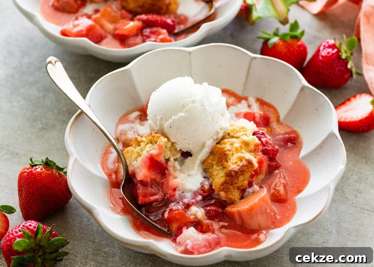 two serving bowls of strawberry rhubarb cobbler topped with ice cream with more strawberries nearby.