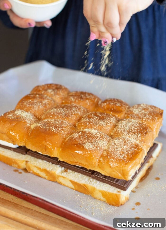 Close-up of a person sprinkling crumbs over a slab of connected bread rolls placed on a baking sheet. Chocolate squares and marshmallow are visible between the layers of bread. A white bowl is partially visible in the background.