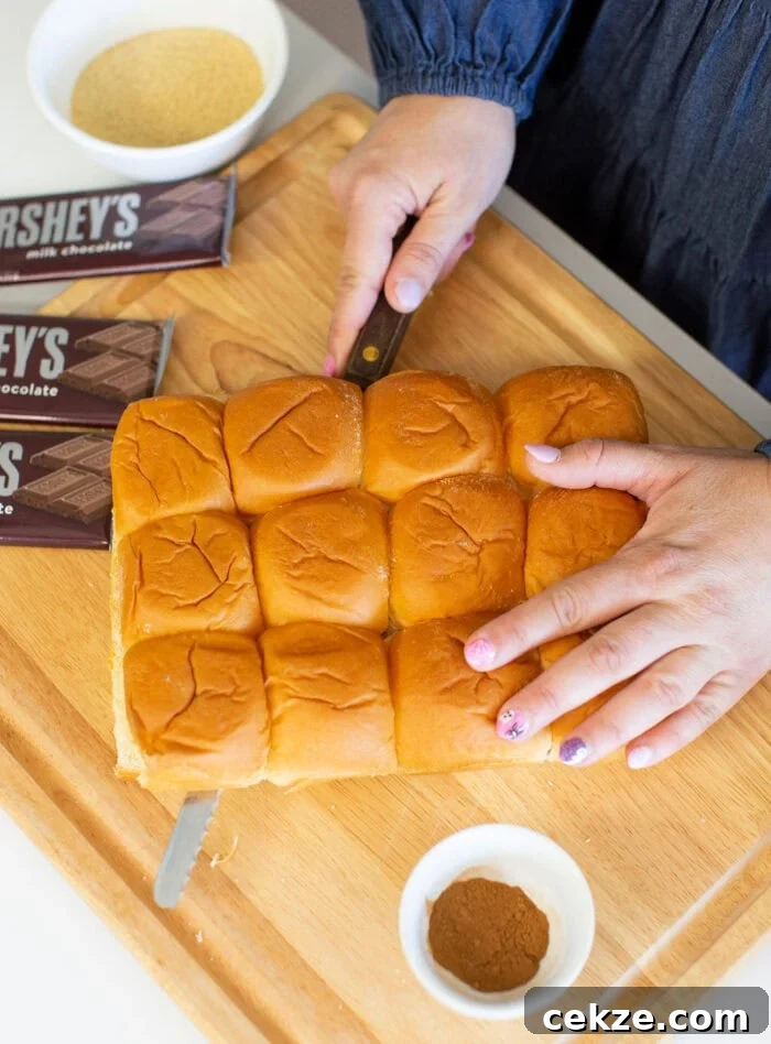 A person slices a tray of small dinner rolls on a wooden cutting board. Two Hersheys milk chocolate bars, a bowl of cinnamon, and breadcrumbs are nearby. The person wears a dark blue top with floral nail art.