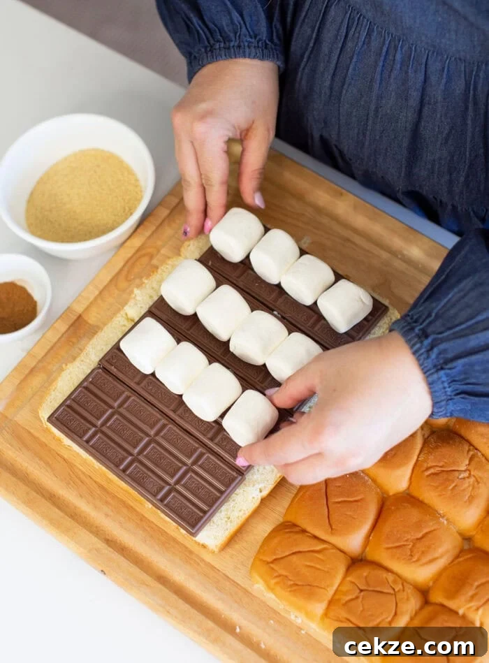 Person assembling a smores dessert with chocolate and marshmallows on a wood board, surrounded by halved bread rolls. Two bowls with brown sugar and cinnamon are nearby.