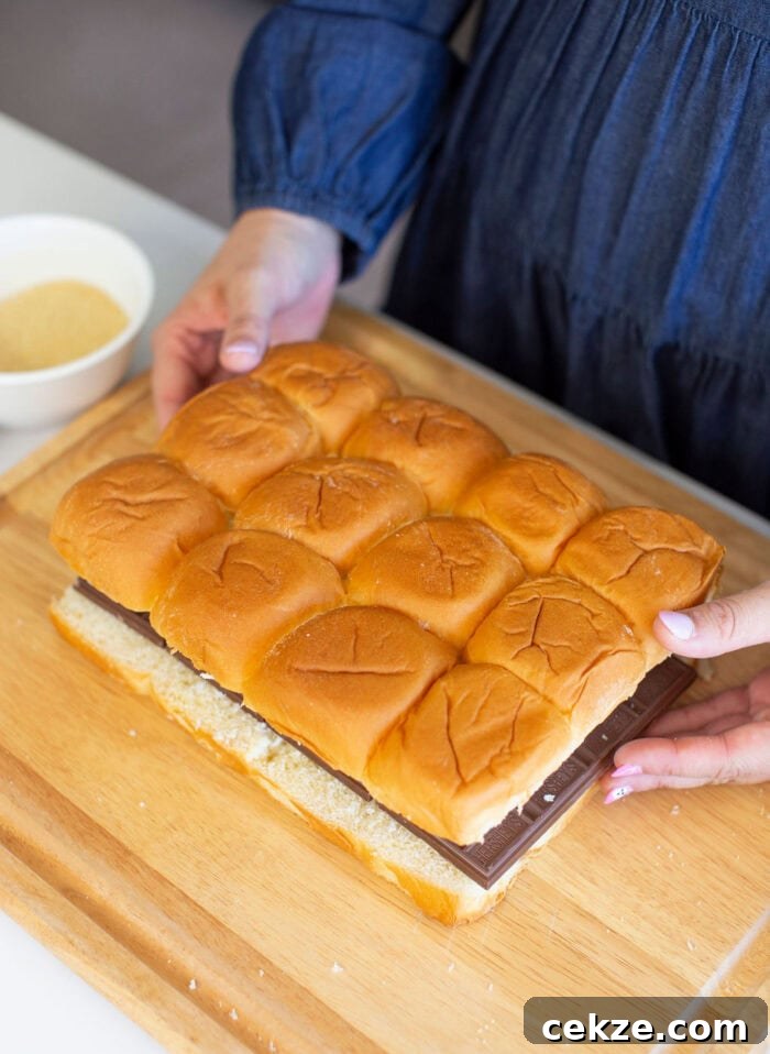 A person is assembling a sandwich with chocolate bars between two layers of soft, golden brown bread rolls on a wooden board. A small bowl with a light-colored ingredient is nearby.