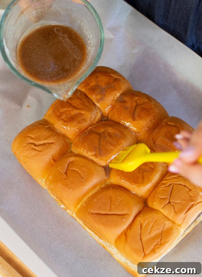 A person is brushing a glaze onto a tray of connected dinner rolls with a yellow brush. A glass container filled with more glaze is nearby on the white parchment paper.