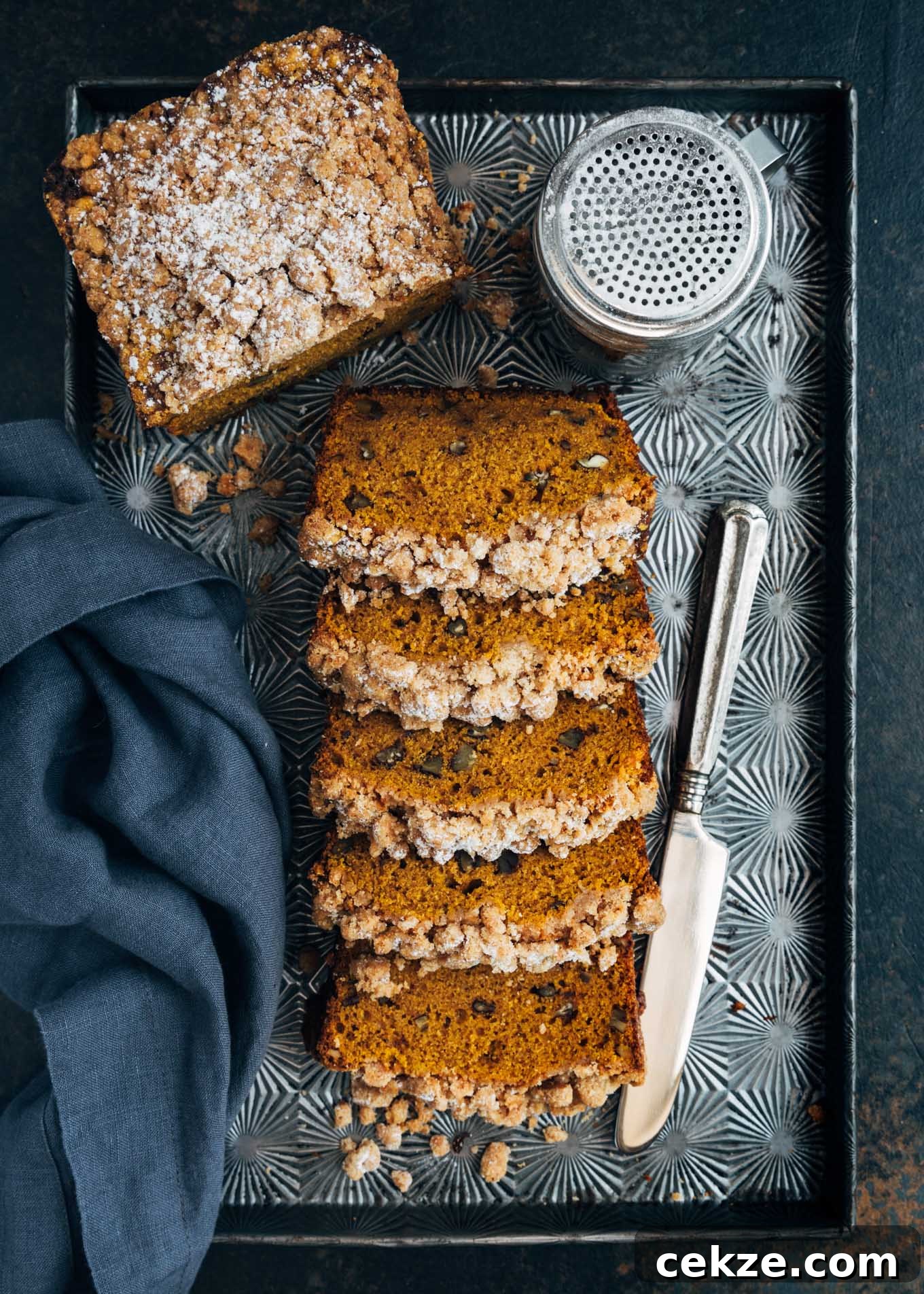 overhead of sliced pumpkin bread with crumb streusel topping on a baking sheet with a silver knife and blue-gray napkin.
