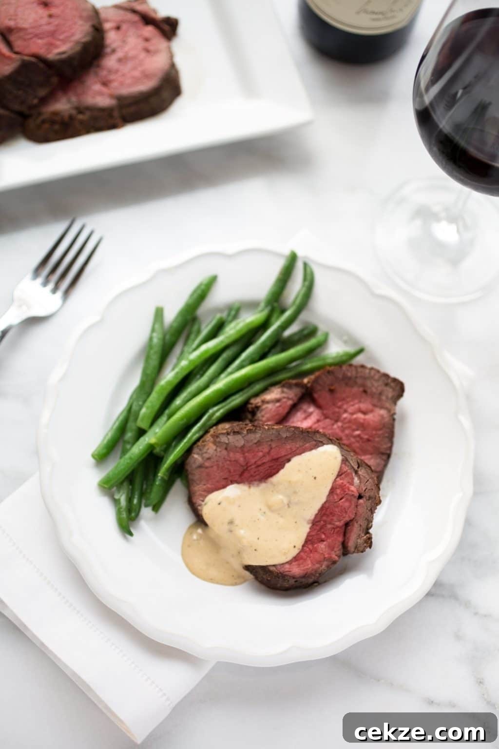 overhead of slices of beef tenderloin on a white plate drizzled with cognac cream sauce, next to cooked green beans.