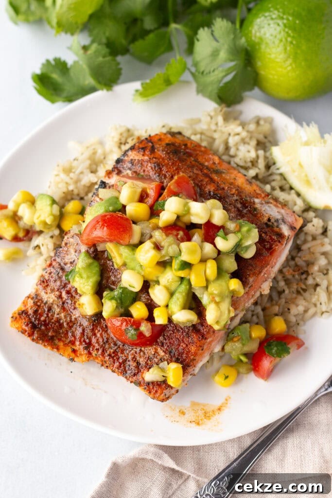 15-Minute Chili Lime Grilled Salmon Recipe 12 Close up of a filet of grilled salmon on a white plate on top of rice with some corn tomato avocado salsa on top of it. A tan cloth napkin is under the plate and a lime and some fresh herbs are next to the plate.