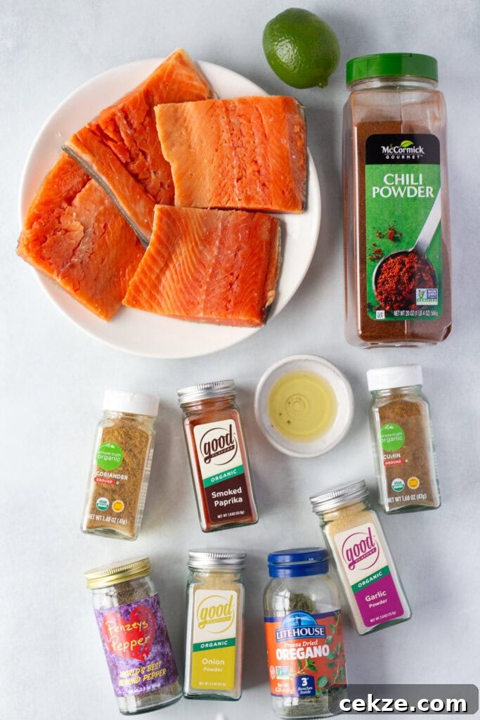 15-Minute Chili Lime Grilled Salmon Recipe 3 Top down shot of ingredients for chili lime grilled salmon on a light gray background, including 4 salmon filets on a white plate, bottles of spices, a lime, and a small gray bowl with oil.