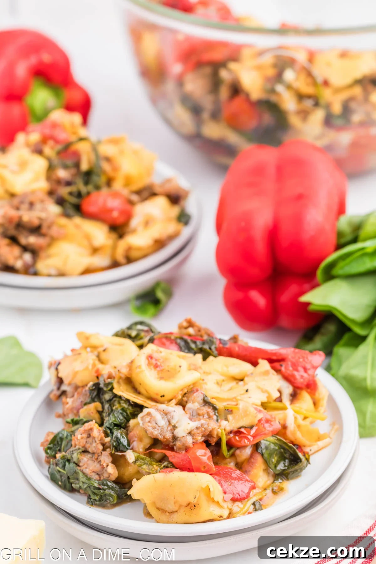 Cooked tortellini pasta mixed with Italian sausage, tomato and spinach on a white plate. Red bell pepper and fresh spinach in the background