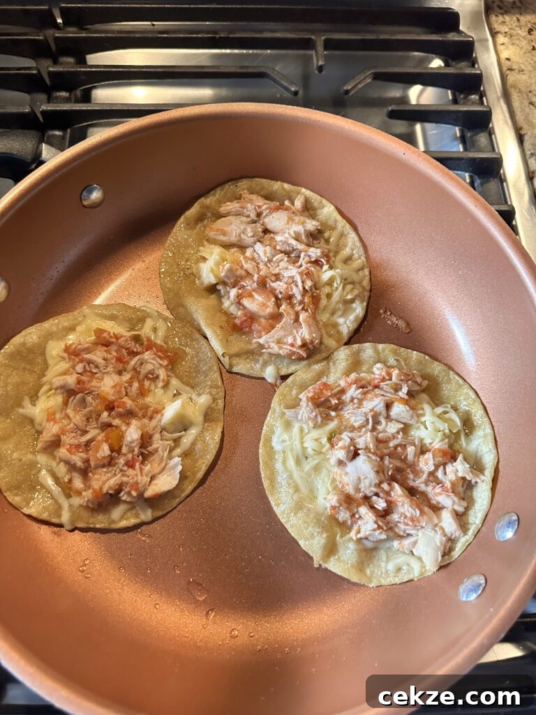Tacos topped with shredded cheese and shredded chicken cooking in an oiled pan