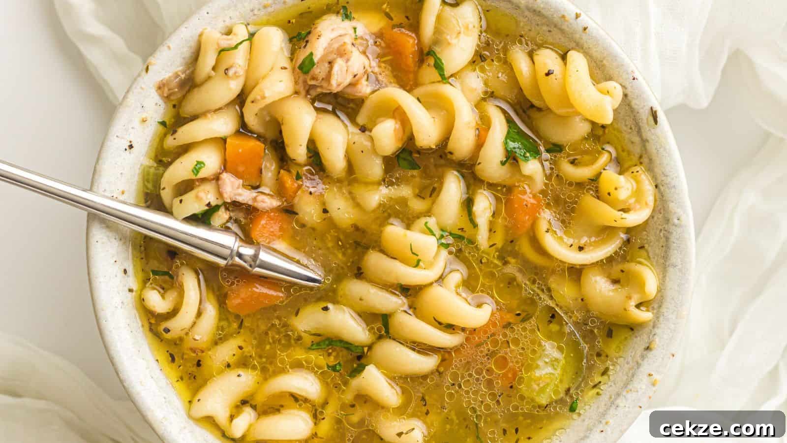 A bowl of rotini pasta soup with chunks of chicken, carrots, and herbs, with a spoon resting inside.