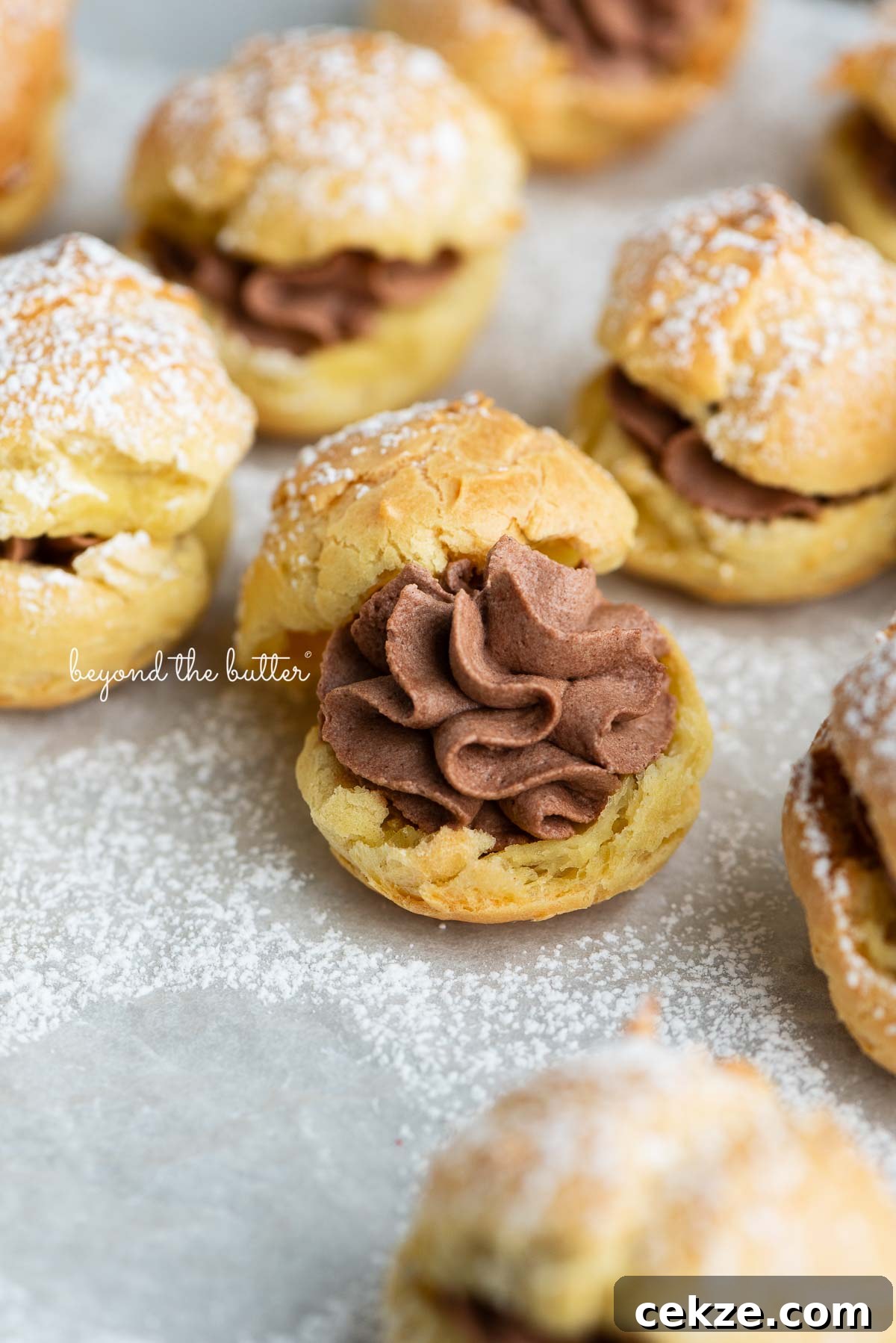 How to Make Classic Cream Puffs: Light Choux Pastry Recipe 2 Cream puffs filled with chocolate whipped cream and dusted with powdered sugar on a parchment paper lined baking sheet.