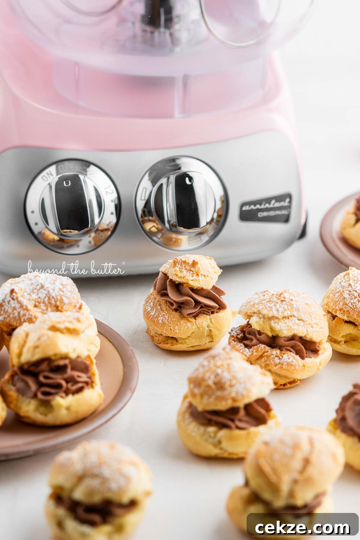 How to Make Classic Cream Puffs: Light Choux Pastry Recipe 3 Cloudy pink Ankarsrum mixer with cream puffs filled with chocolate whipped cream and dusted with powdered sugar placed around it on pink dessert plates and white marbled background.