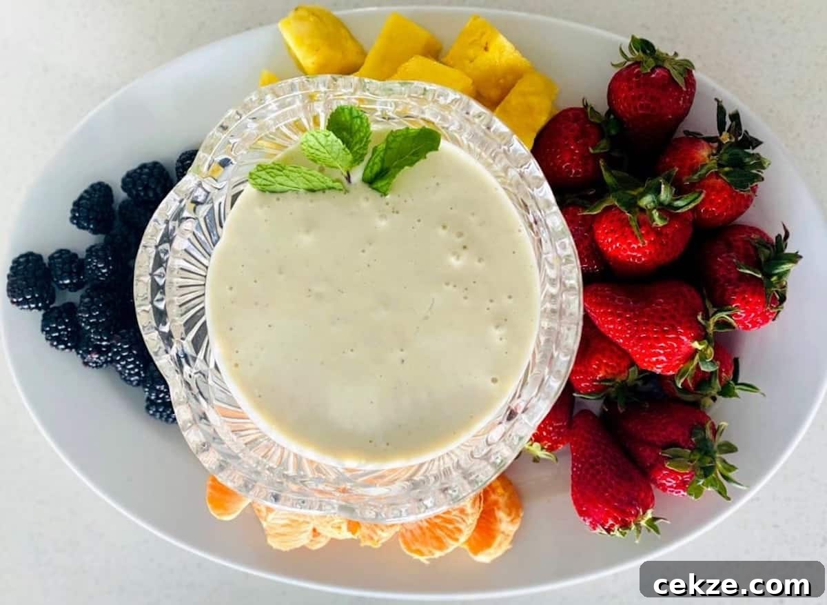 Vegan fruit dip recipe in a bowl garnished with a mint leaf on a plate with strawberries, mandarin oranges, pineapple, and blackberries.
