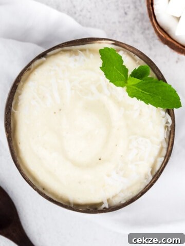 A picture of a white smoothie inside a coconut bowl, garnished with fresh mint and coconut flakes. There is a white linen cloth, fresh coconut, and part of a wooden spoon shown decoratively in the background.