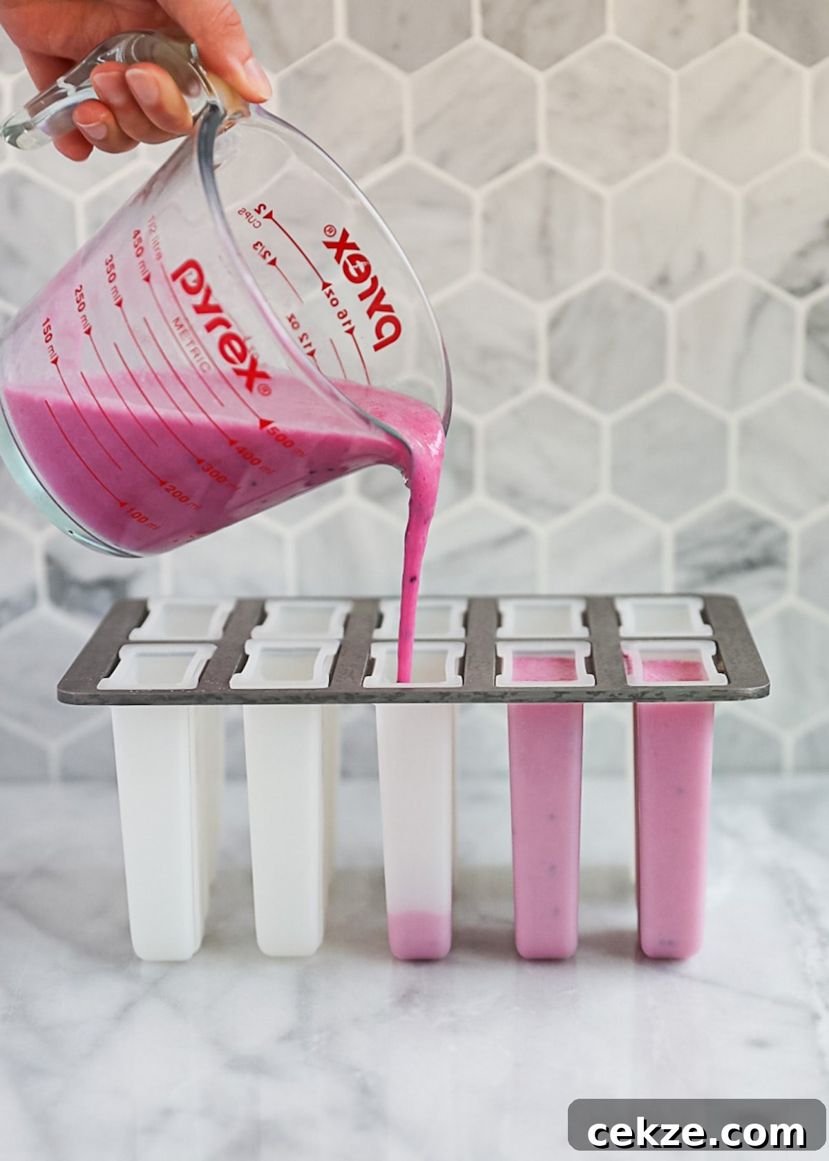 Refreshing Dragon Fruit Popsicle Recipe for Summer Treats 5 A hand holding a measuring cup that is pouring the unfrozen pink popsicles into popsicle molds.
