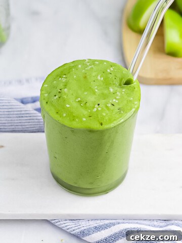 Green smoothie in a clear cup, garnished with hemp seeds, with a clear glass straw placed in the smoothie. It's sitting on a white marble tray that is on a blue and white striped dish towel. There are sliced green apples in the background.