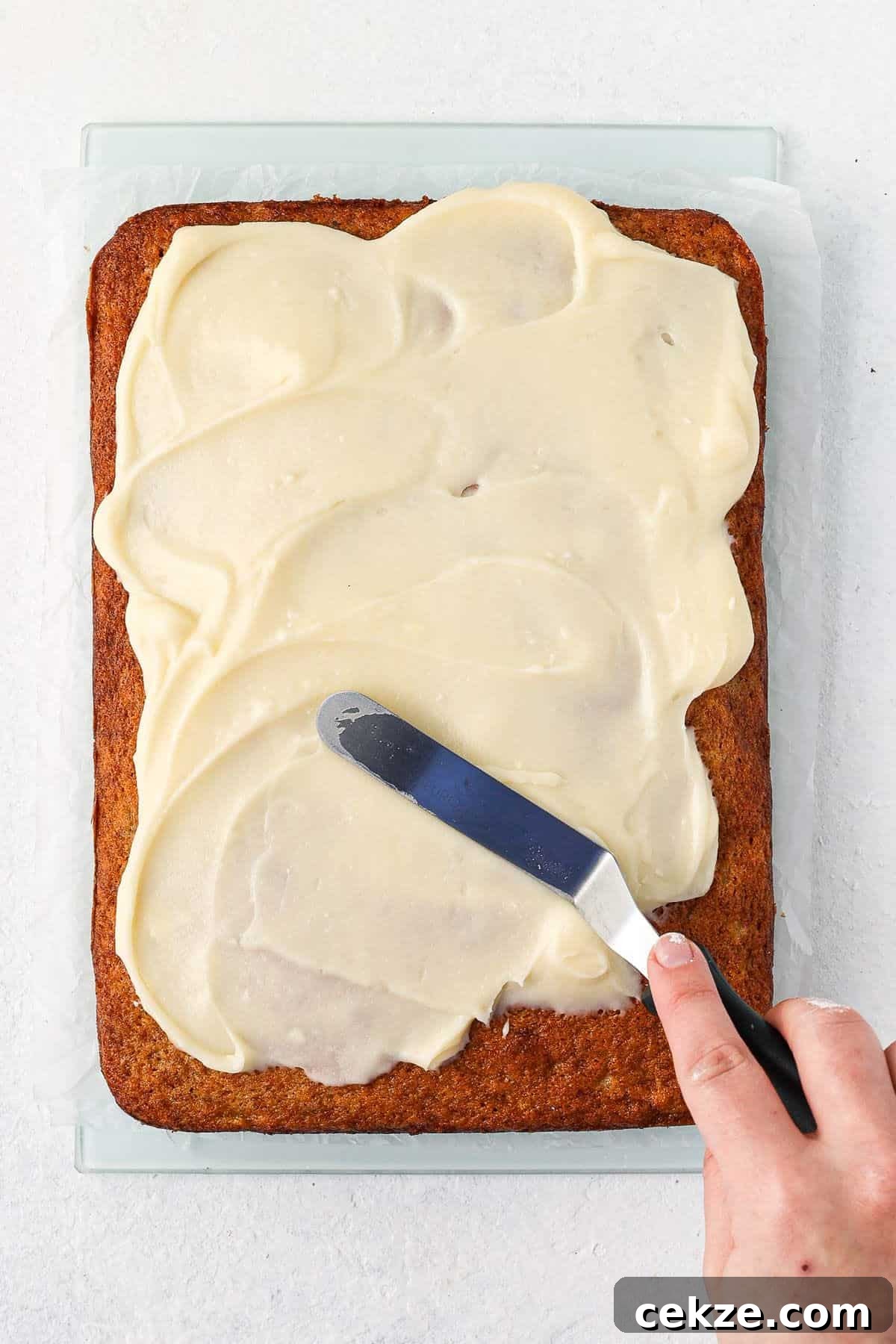 Banana Slice being iced with cream cheese frosting, showing a hand with a palette knife smoothing frosting.