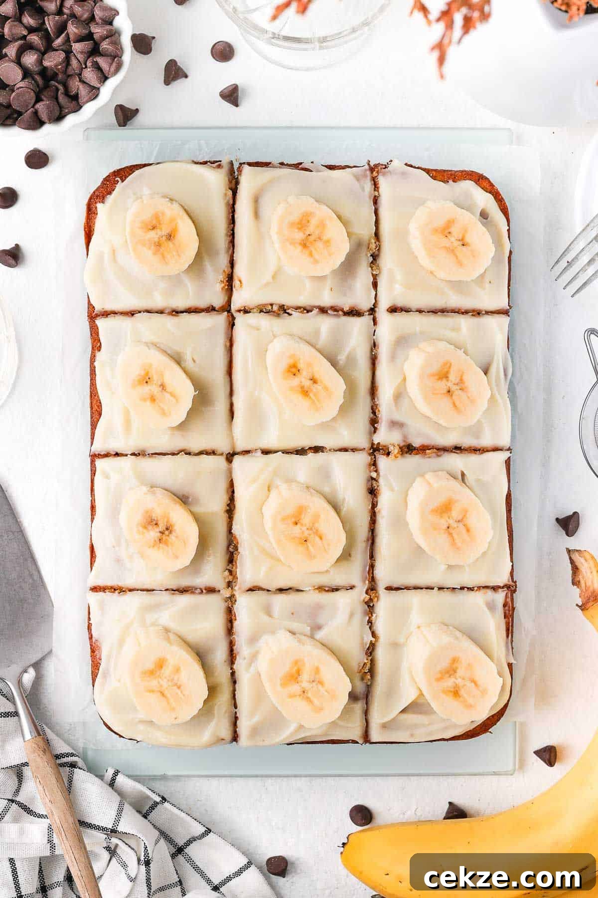Banana Slice with cream cheese frosting, cut into 12 squares and topped with a slice of fresh banana, sitting on a glass board.