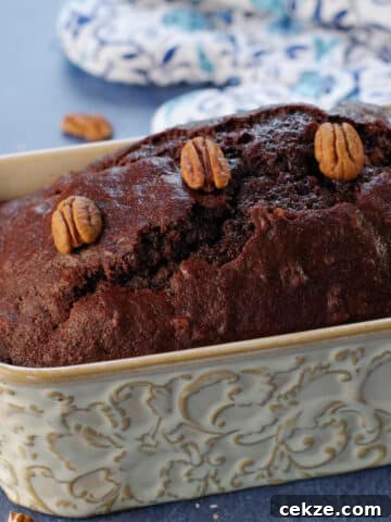 Entire loaf of chocolate zucchini loaf with blue patterned oven mitts in the background and pecans strewn about