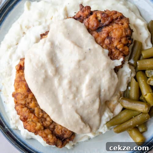 piece of chicken fried steak with white gravy on top of mashed potatoes with green beans on the side.