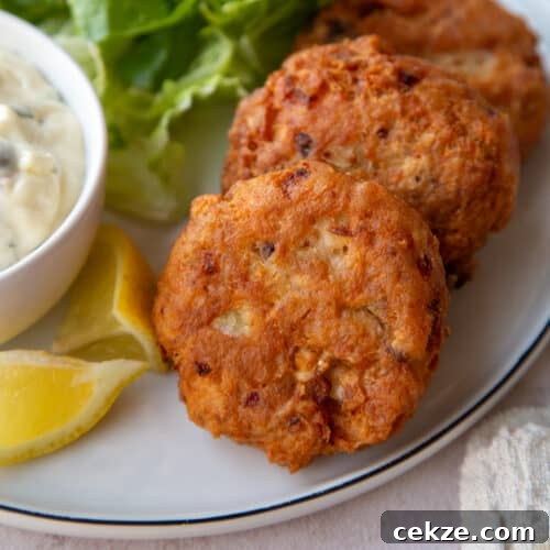 2 fried Salmon Patties on a dinner plate next to Tartar Sauce and lemon wedges.