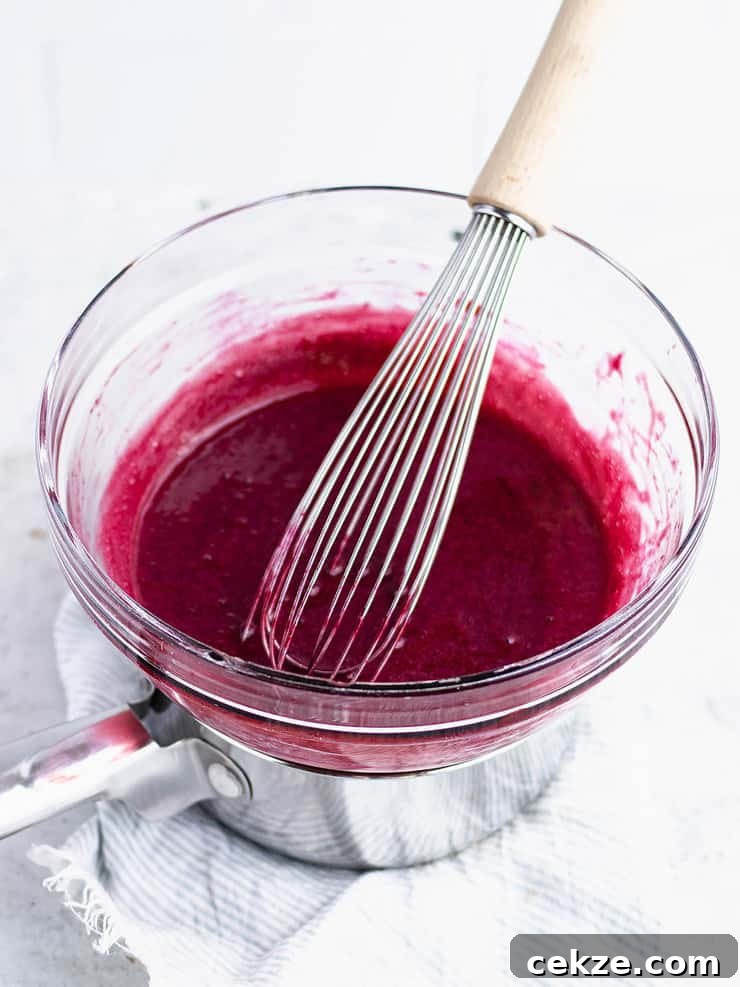 Blackberry curd in a clear glass bowl over a pot of simmering water with a wooden handled whisk leaning against the righthand side.