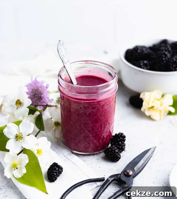 Jar of blackberry curd with a silver spoon sticking out of the top. Berries, flowers and a pair of floral scissors are laid out around it.