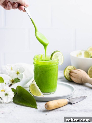 Spoon with lime curd dripping from it back into a glass mason jar. White flowers, a knife and limes are in the background.