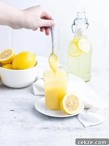 Lemon curd dripping from a silver spoon into a ½ pint jar of curd. Lemons in a bowl and lemonade in a decanter in the background.