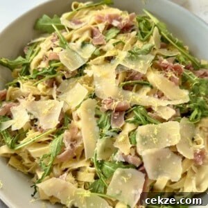 A bowl of tagliatelle pasta topped with arugula, thinly sliced prosciutto, and shaved Parmesan cheese. The dish is garnished with lemon zest, creating a fresh and colorful appearance. A cup is partially visible in the background.