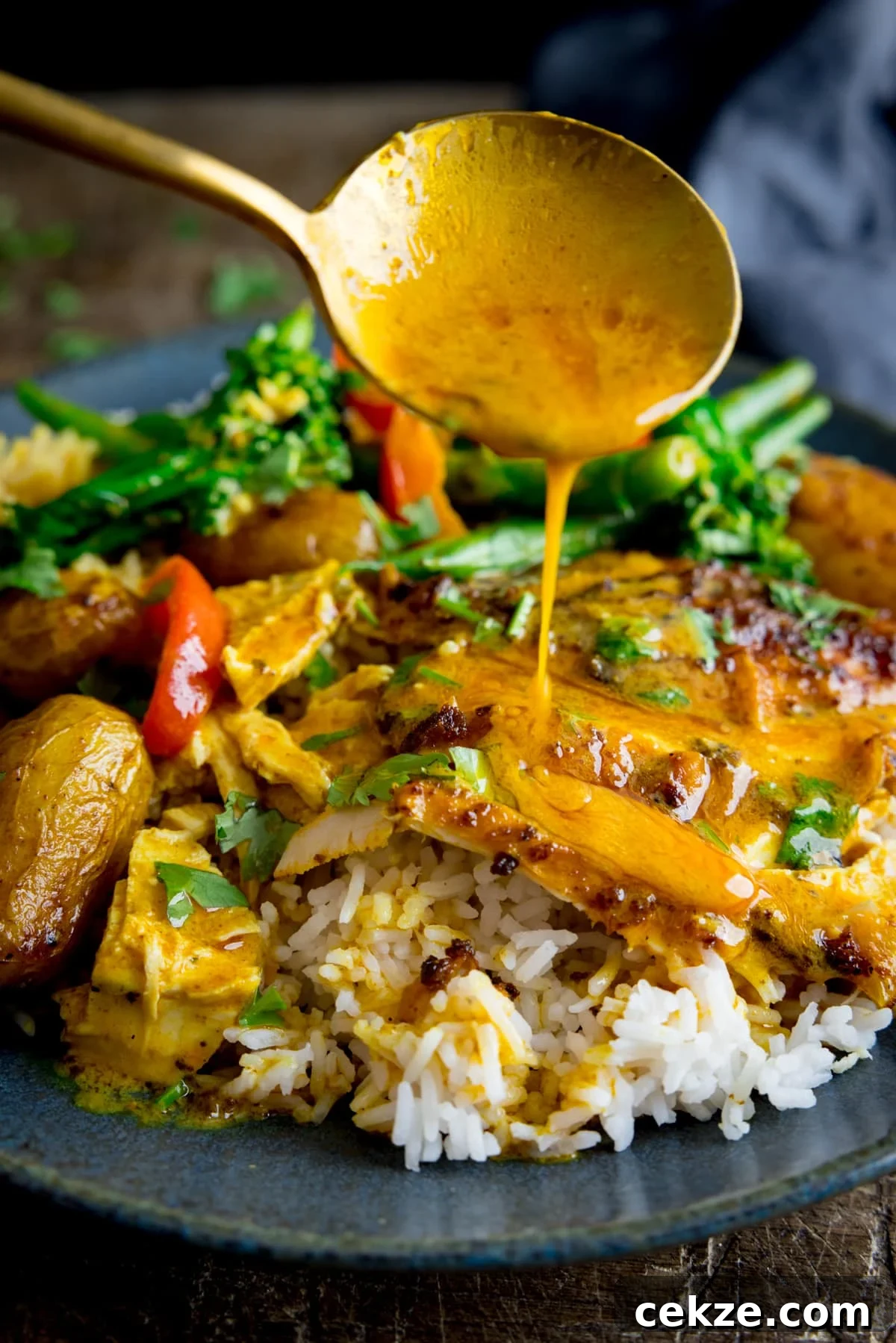 Curry-Roasted Chicken with Crispy Skin and Aromatic Spices 11 Curry sauce being spooned onto slices of curry roast chicken which is on a plate with rice and vegetables.