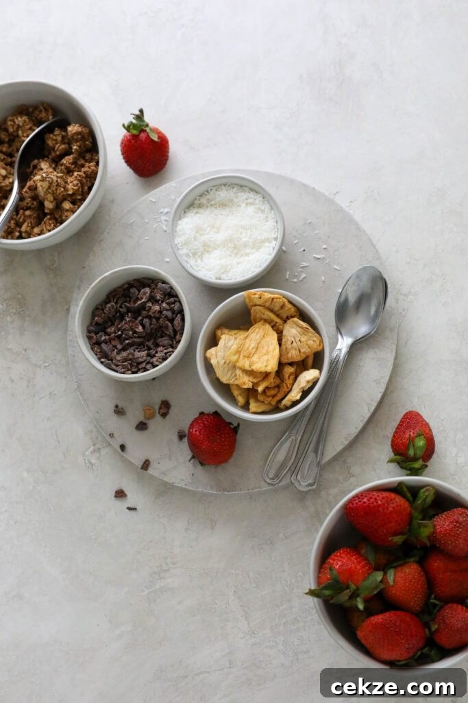 Strawberry Mango Smoothie Bowl Recipe for Creamy Tropical Breakfast 6 granola, cacao nibs, shredded coconut, and dried pineapple on circle with two spoons and fresh strawberries
