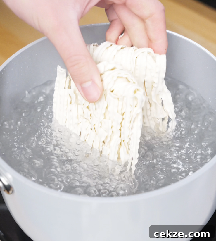 A hand dropping a block of instant noodles into a pot of rapidly boiling water, marking the start of the noodle prep for chili garlic butter noodles.