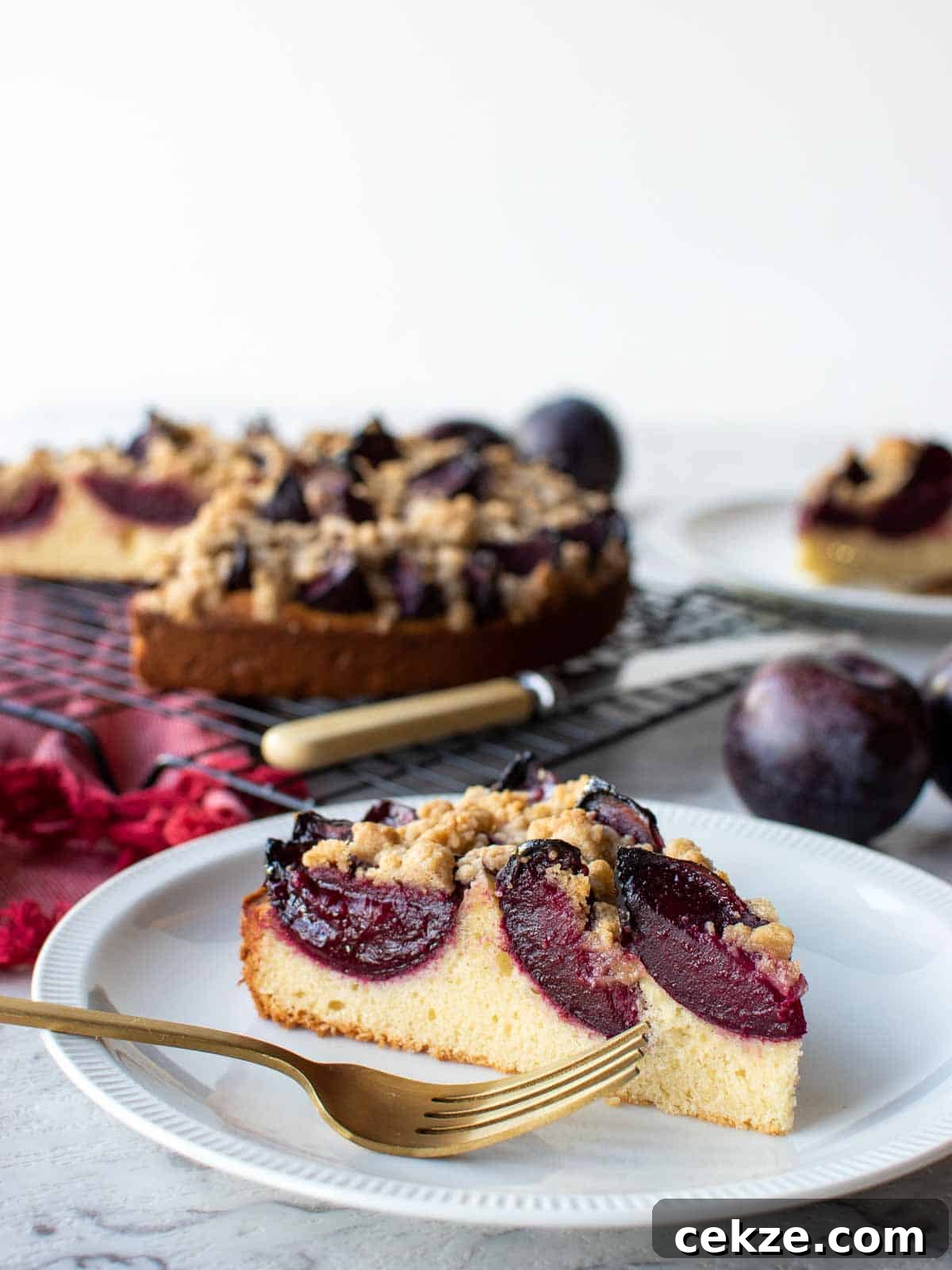 Classic German Plum Cake Recipe (Zwetschgenkuchen) 11 A slice of plum cake on white plate with whole cake in the background.