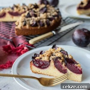 Classic German Plum Cake Recipe (Zwetschgenkuchen) 12 Wedge of plum cake on white plate with gold fork and whole cake in the background.