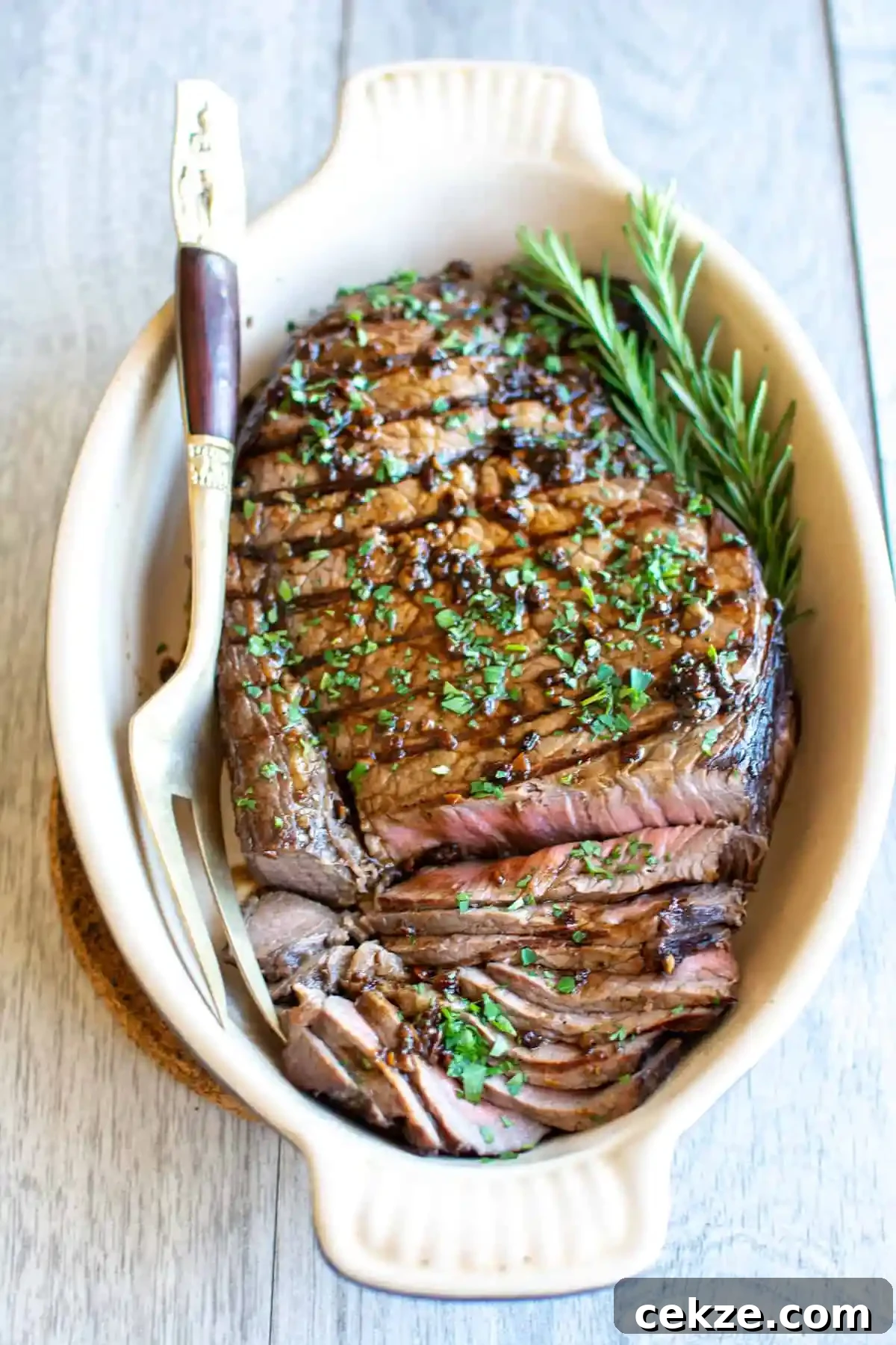 A white, oval platter filled with a grilled London Broil steak topped with chopped parsley and a carving fork next to it. 