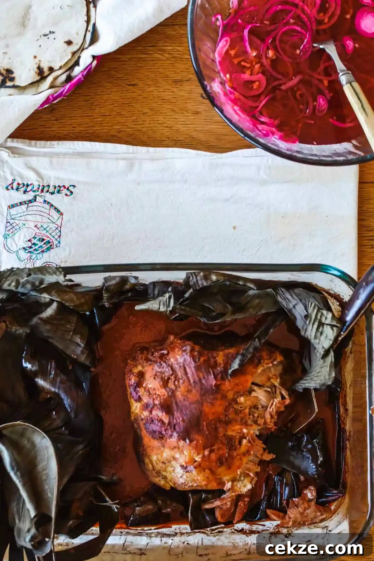A glass dish with a charred banana leaf and a Cochinita Pibil roast next to a bowl of pickled onions sitting next to a white cloth. 