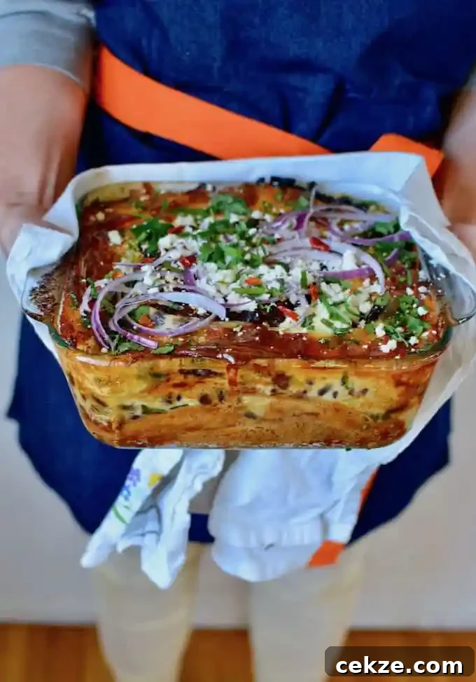 A woman in a blue apron with an orange belt holding a casserole dish of baked enchiladas topped with red onion slices. 