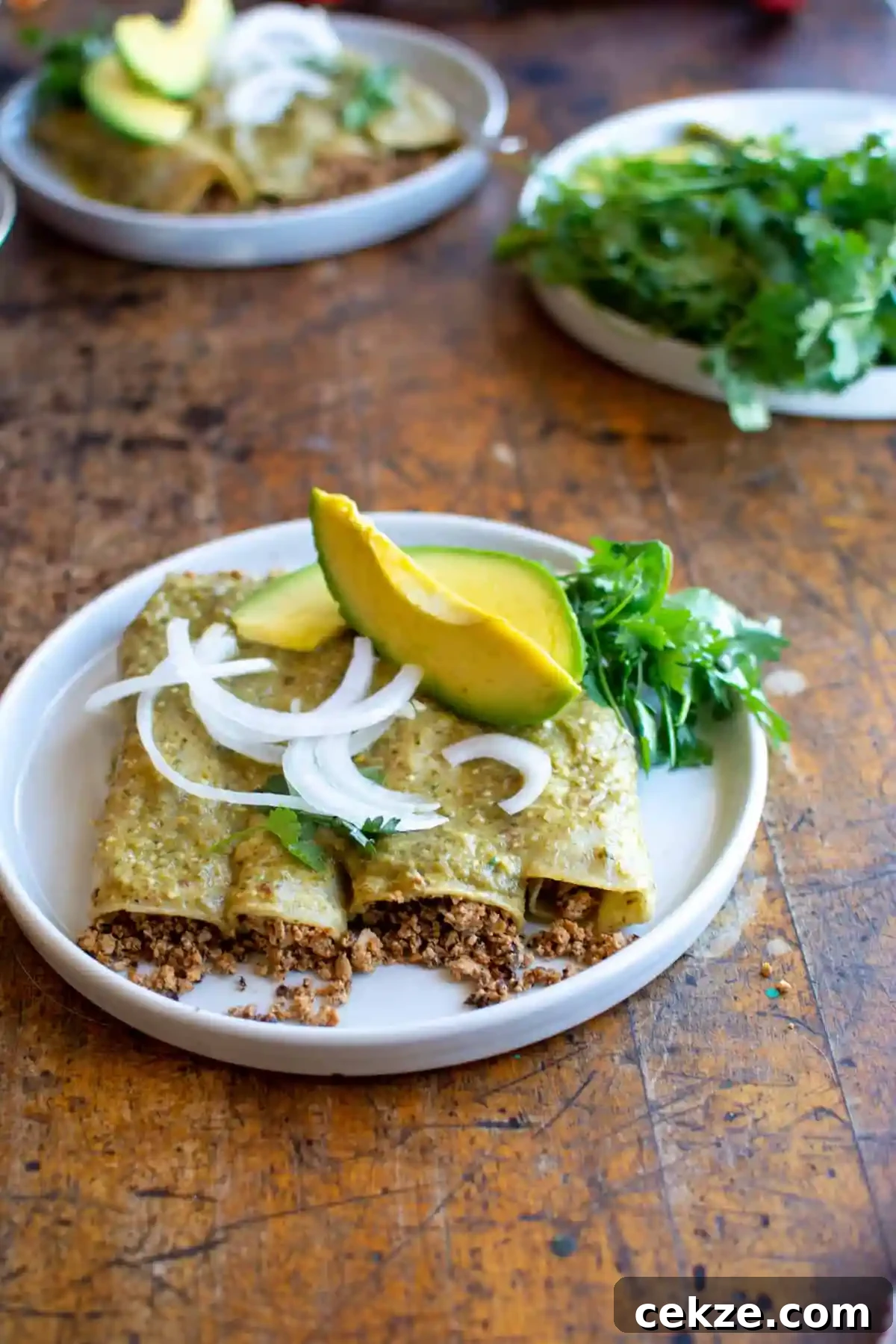 A white plate on a wood table. The plate has four vegan enchiladas on it and is topped with white onion slices and two wedges of avocado. 