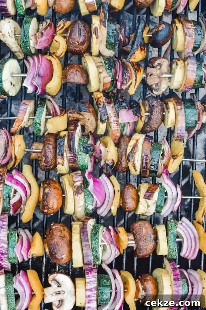 A grill full of vegetable skewers. Skewers of sliced zucchini, red onion, yellow pepper, mushrooms, all on metal skewers. 