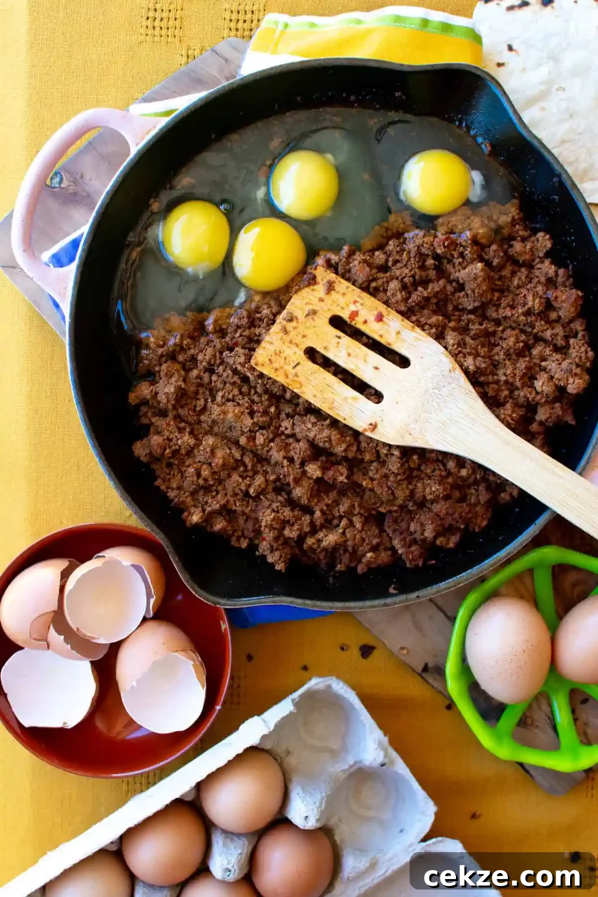 A frying pan with chorizo and eggs in it that is sitting on a yellow table cloth with a wooden spatula in it. With a bowl off to the side. 