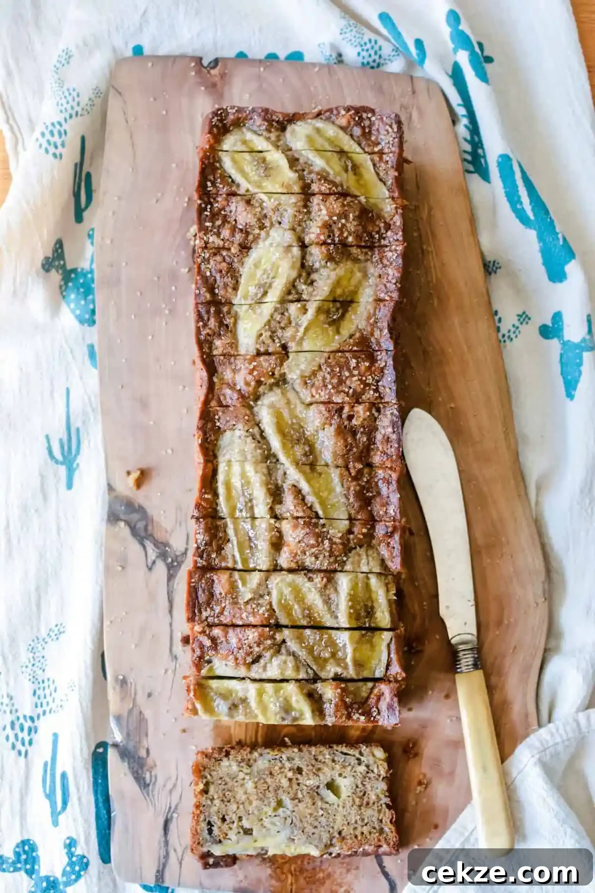 A long loaf of banana bread topped with bananas and cut into thick slices sitting on a wood board with a butter knife next to it. 