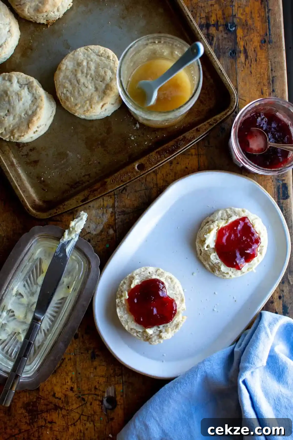 An oval plate with a biscuit cut in half and spread with butter and jam with a blue kitchen towel next to it and a butter dish. 