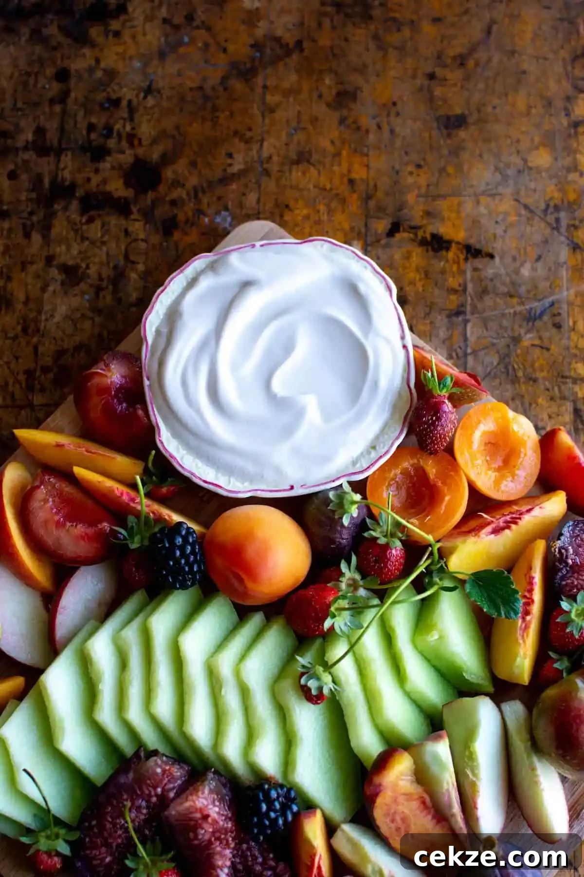 A platter with sliced and cut fruit on it and a small bowl of sour cream fruit dip in one corner on a wood table. 