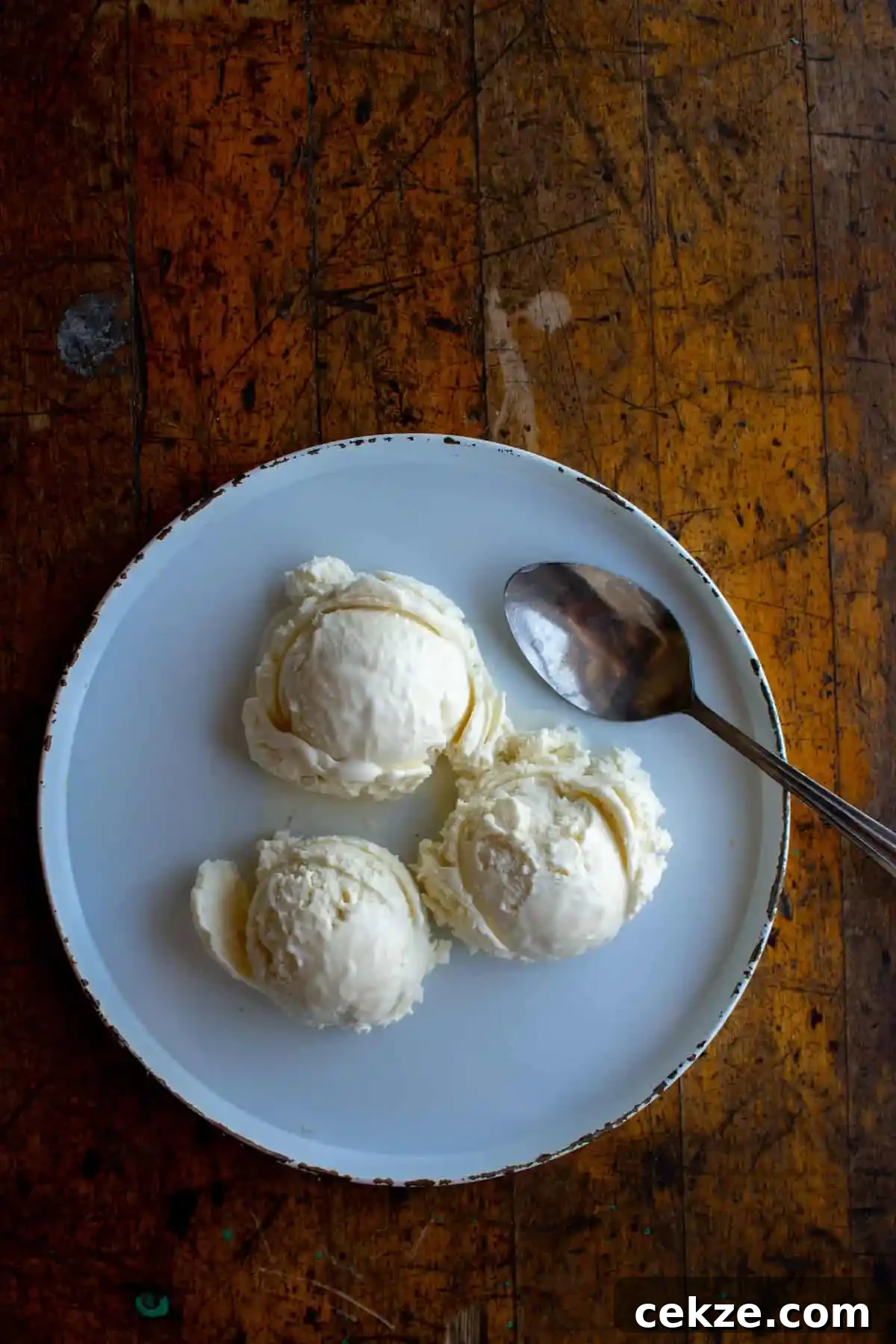 A wood table with a white plate on it and three scoops of vanilla ice cream on top of the plate with a spoon sitting to one side. 