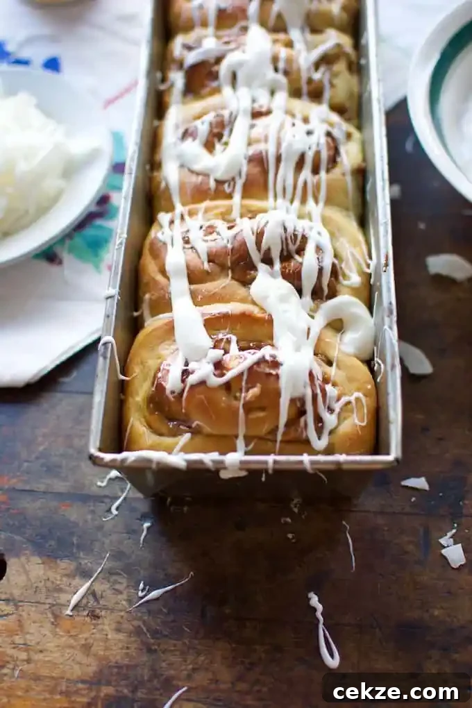 A long rectangular pan of sweet rolls drizzled with coconut frosting on a wood table with a white tablecloth and a plate. 