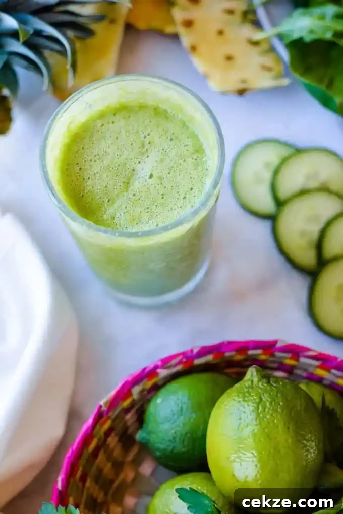 A tall glass with green Jugo Verde in it sitting on a white table with slices of cucumber next to it and pineapple behind it. 