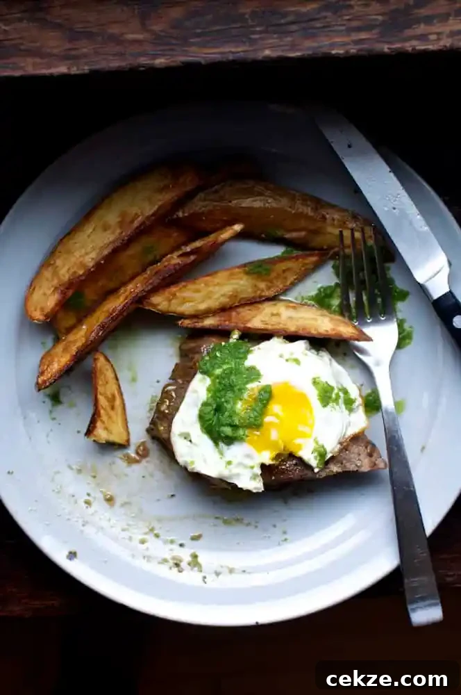 A gray plate on a wood counter with a piece of steak topped with a sunny side up egg and green salsa next to some steak fries. 