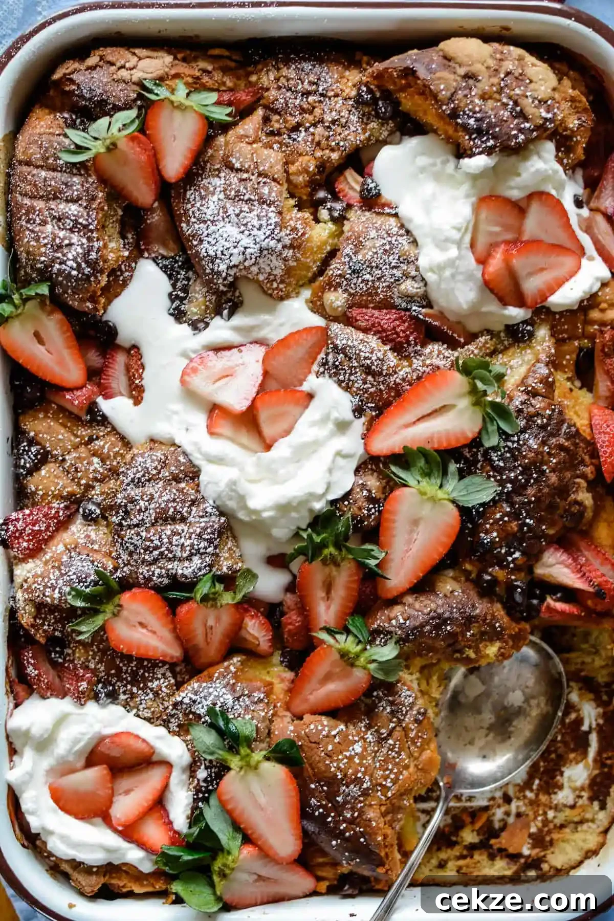A pan of tres leches French toast casserole topped with whipped cream and strawberries and a piece taken out of one end. 